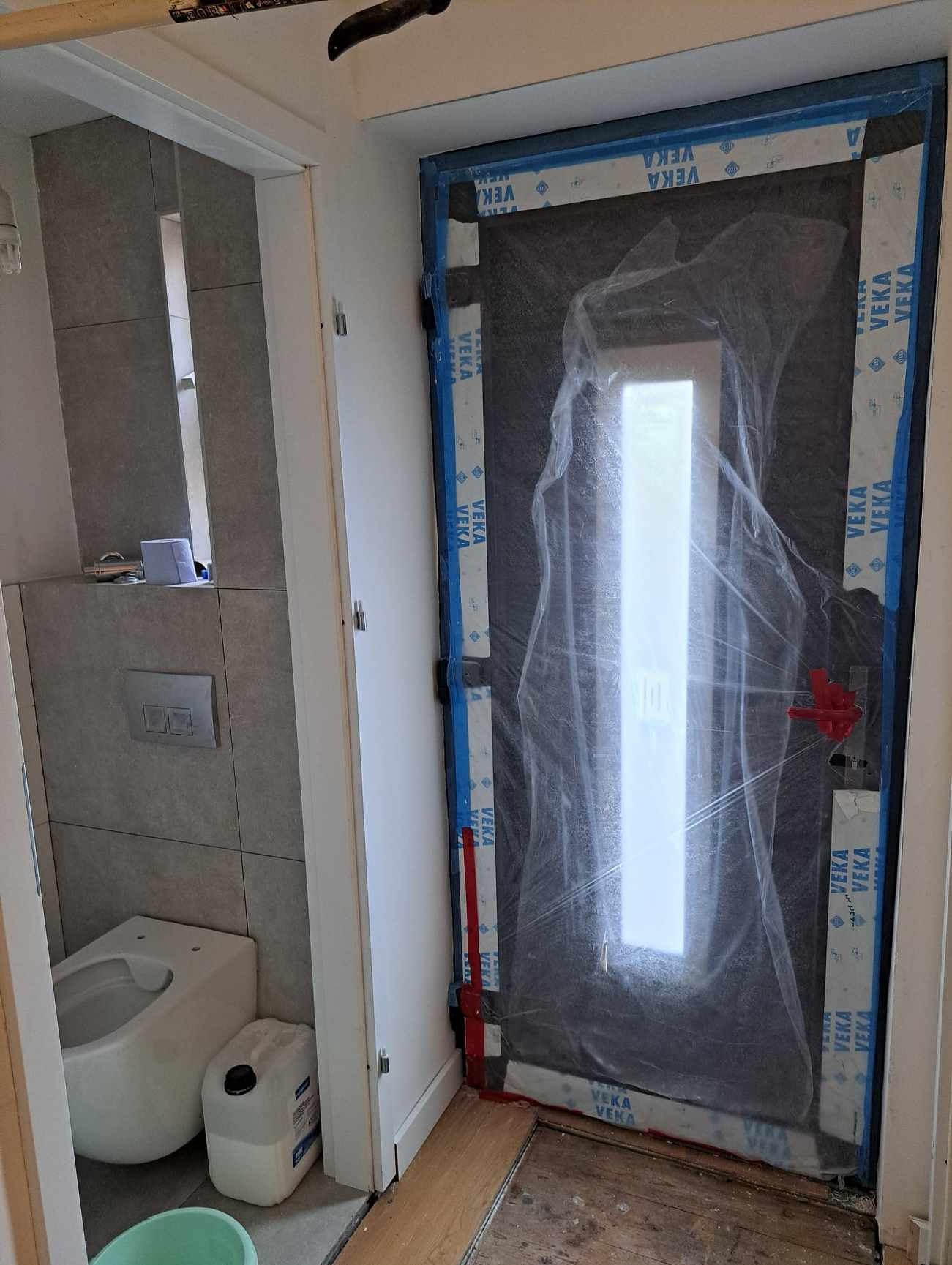Nowe drzwi wejściowe do łazienki, zabezpieczone folią ochronną z logo VEKA, widoczne w trakcie remontu mieszkania, z fragmentem toalety w tle.