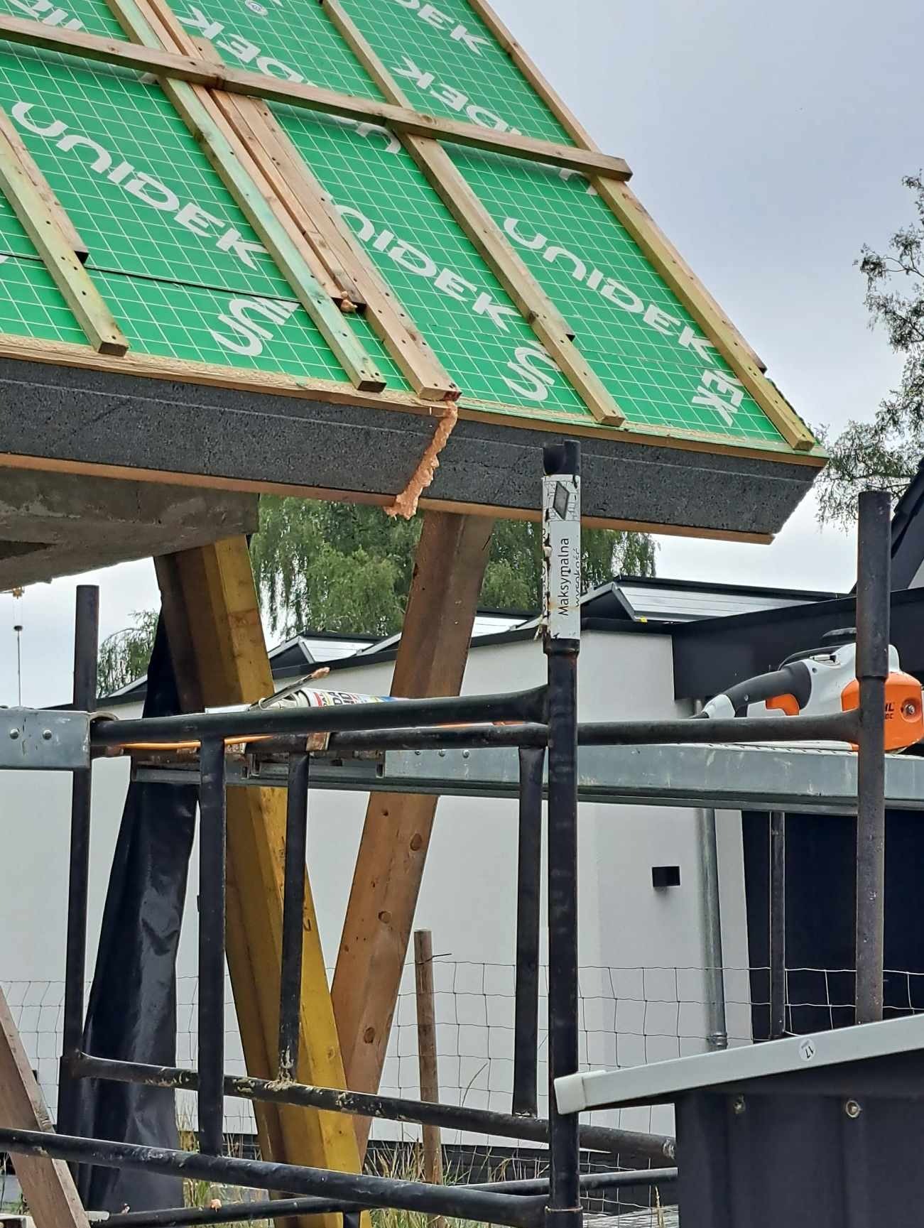 Fragment dachu w trakcie budowy z widocznym deskowaniem, membraną UNIDEK, izolacją termiczną i elementami konstrukcyjnymi, widziany z poziomu rusztowania.