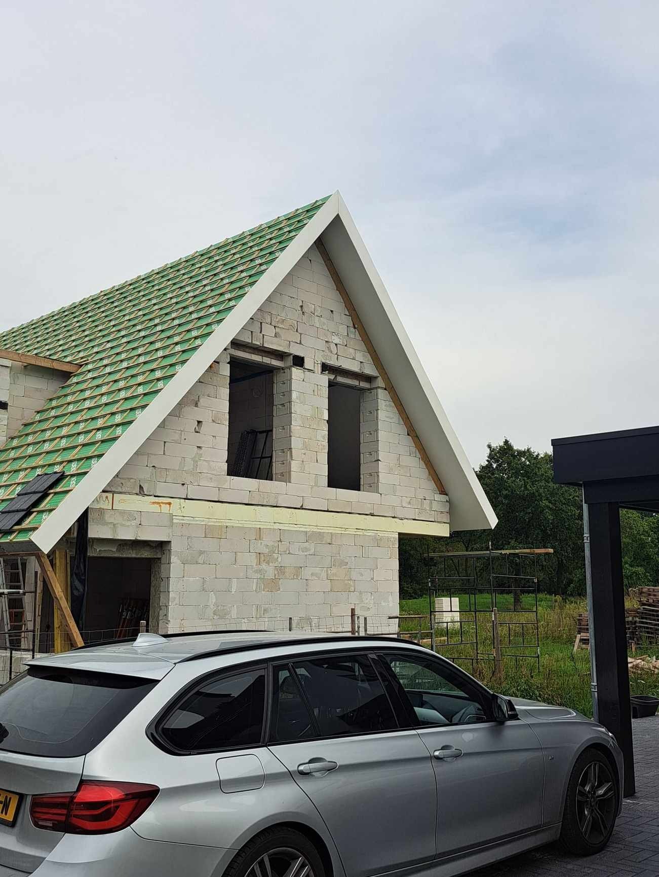 Surowy stan domu w trakcie budowy z widocznym zielonym dachem, szarym samochodem osobowym na pierwszym planie i rusztowaniami w tle.