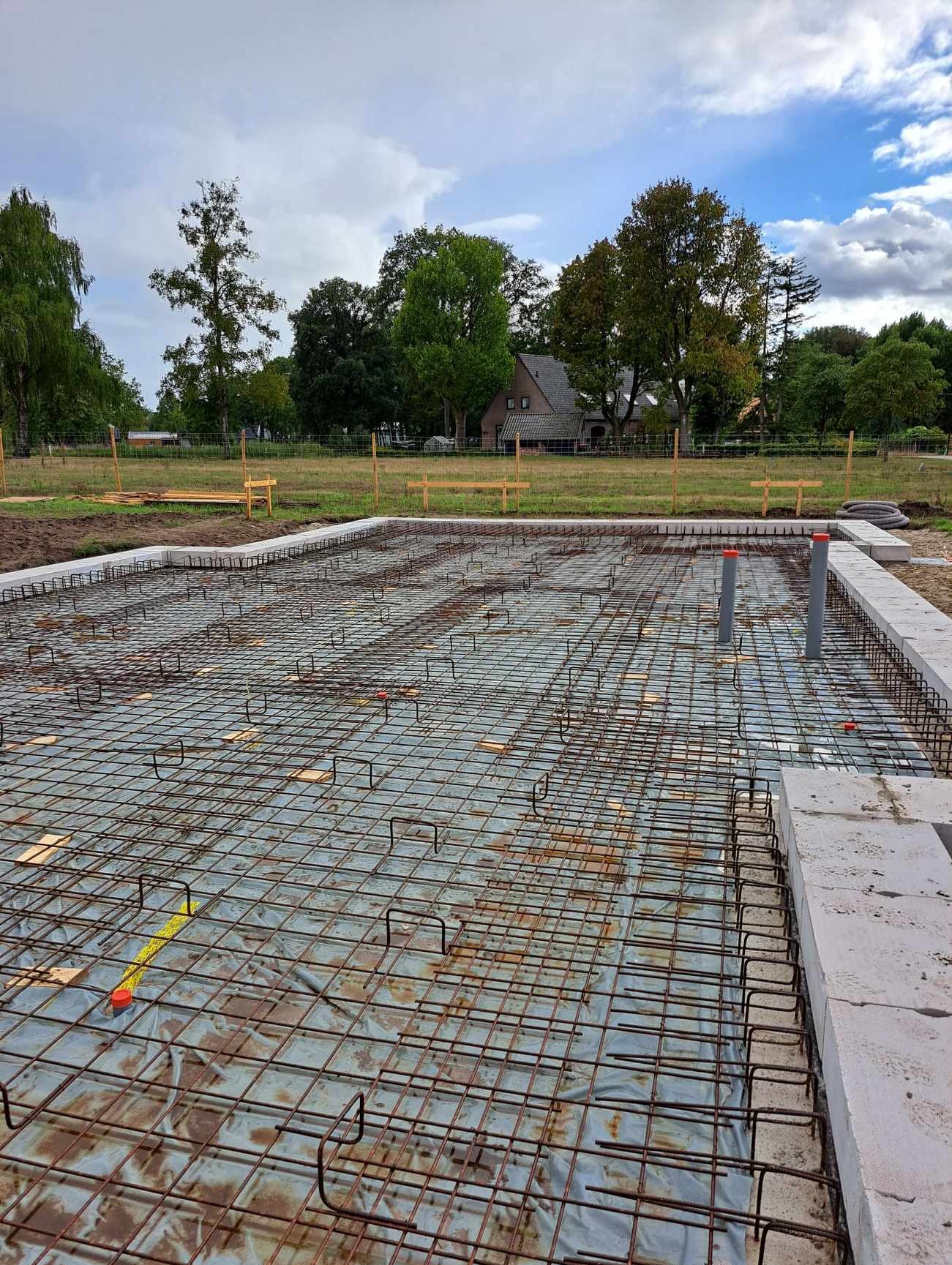 Przygotowanie zbrojenia płyty fundamentowej pod budynek jednorodzinny na działce z widokiem na dom i drzewa.