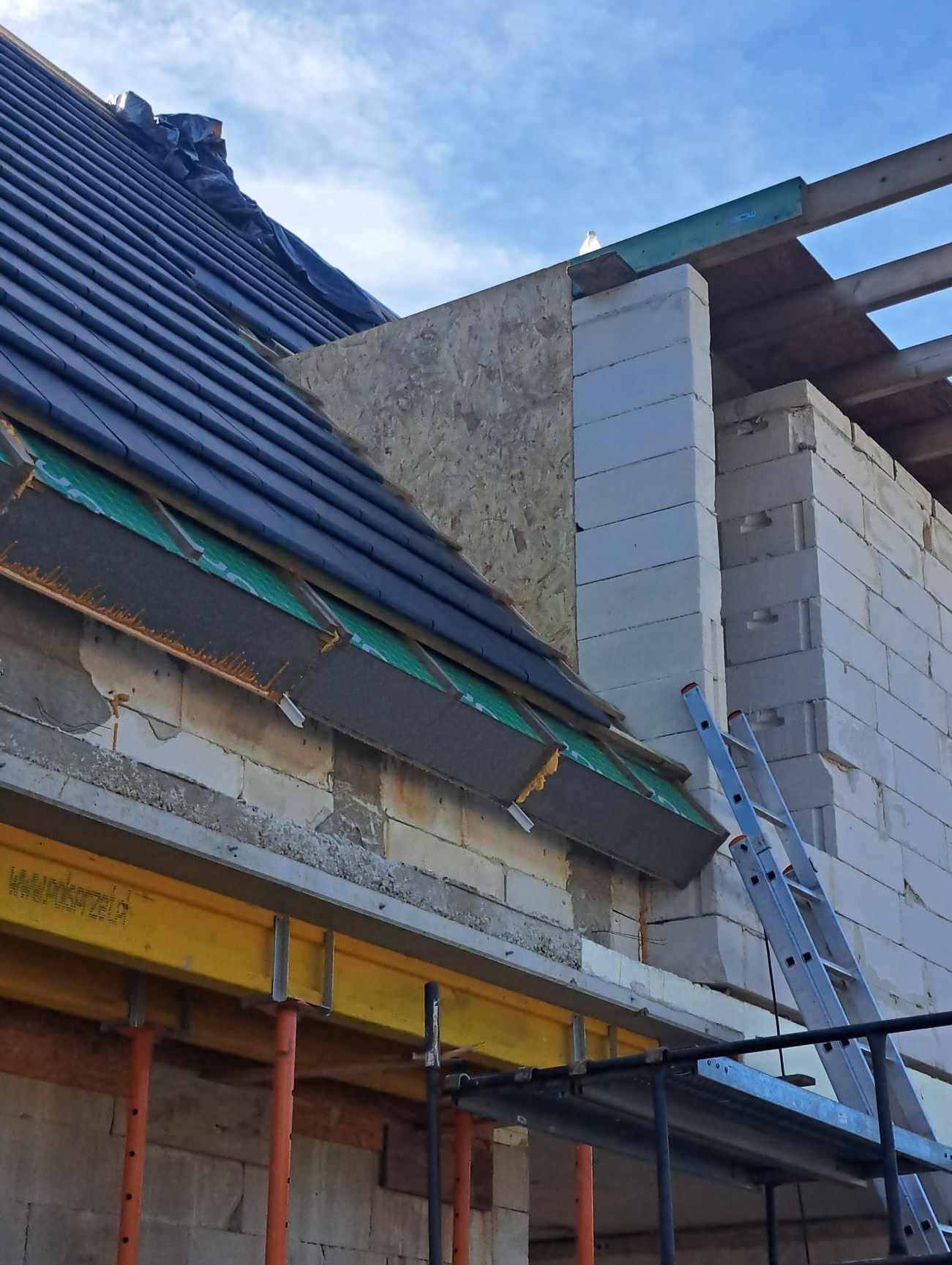 Fragment dachu w trakcie budowy, widoczne warstwy izolacyjne, dachówka, murowane ściany z białych bloczków, rusztowanie i drabina.