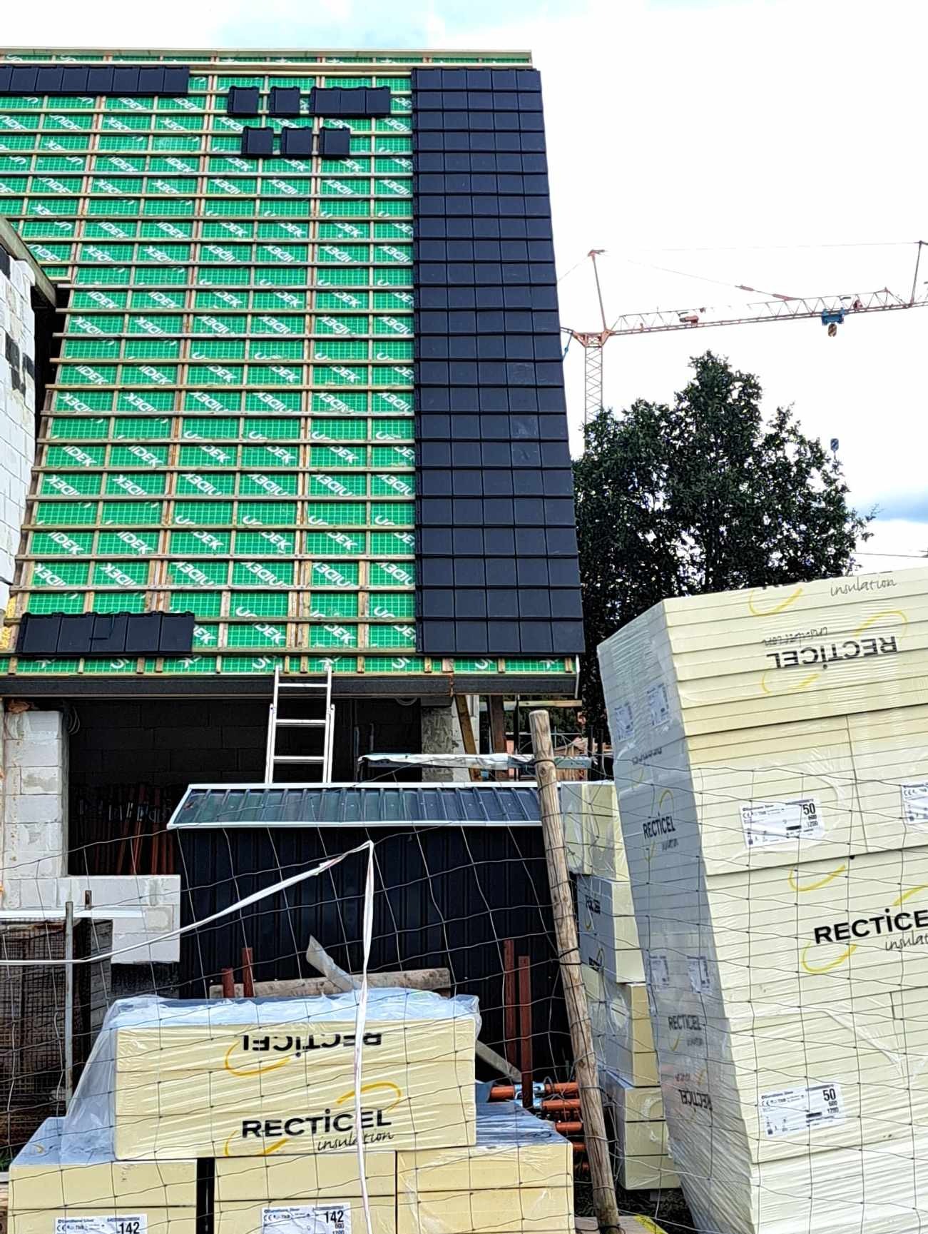 Fragment dachu w trakcie układania czarnej dachówki, widoczna konstrukcja z zieloną membraną, w tle żuraw budowlany i stosy izolacji Recticel.