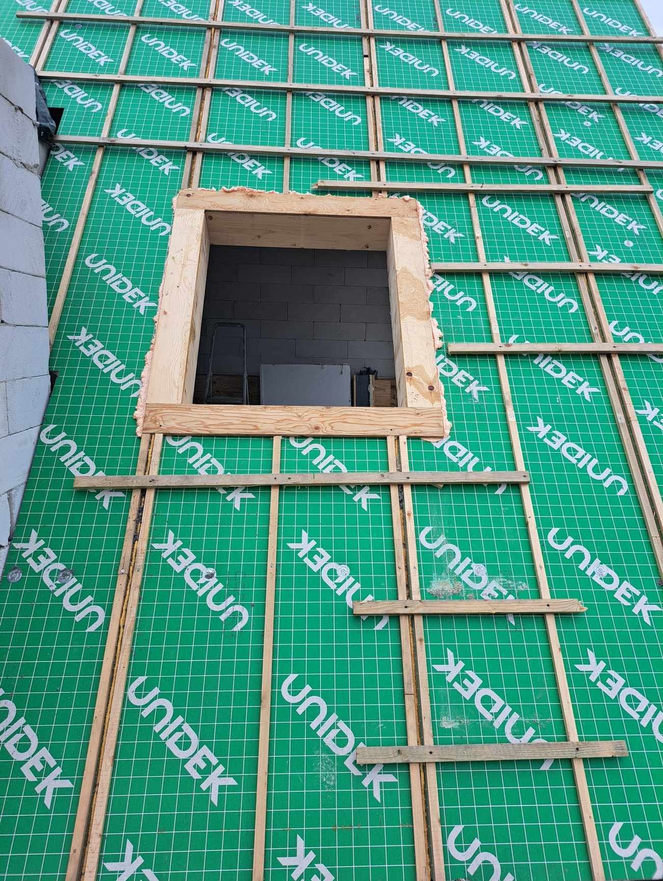 Konstrukcja dachu z oknem dachowym w trakcie budowy, widoczne łaty, folia paroizolacyjna z nadrukiem UNIDEK oraz fragment ściany z pustaków.