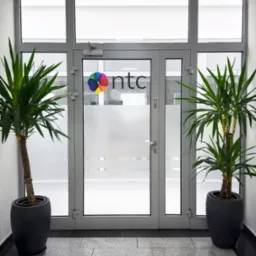 Szklane drzwi wejściowe do biura z logo firmy NTC i numerem 14, flankowane przez dwie juki w ciemnych donicach na szarej posadzce.