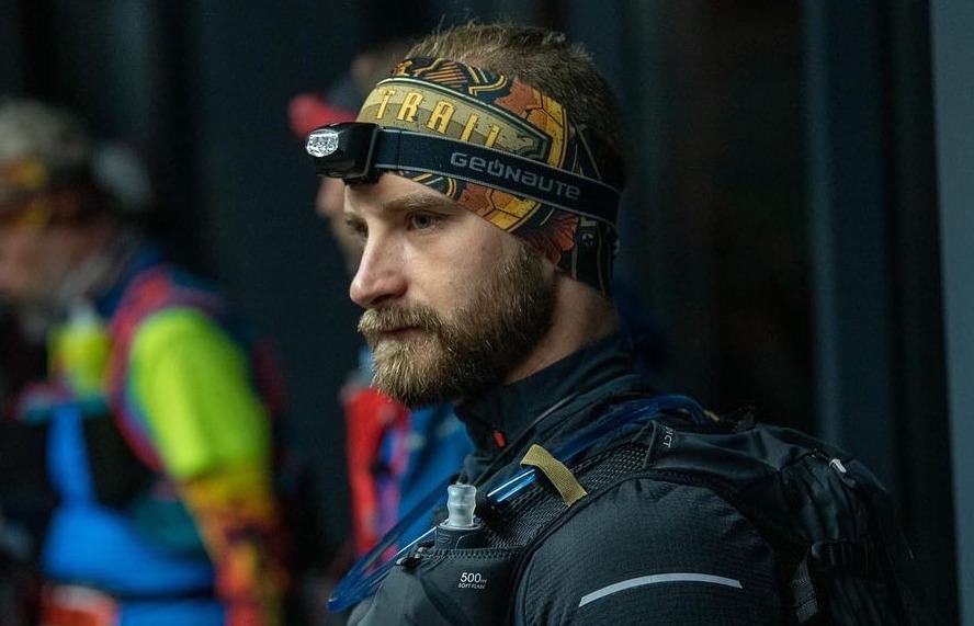 Mężczyzna z brodą w opasce z czołówką, ubrany w strój do biegania z plecakiem z bukłakiem, w tle inni biegacze.