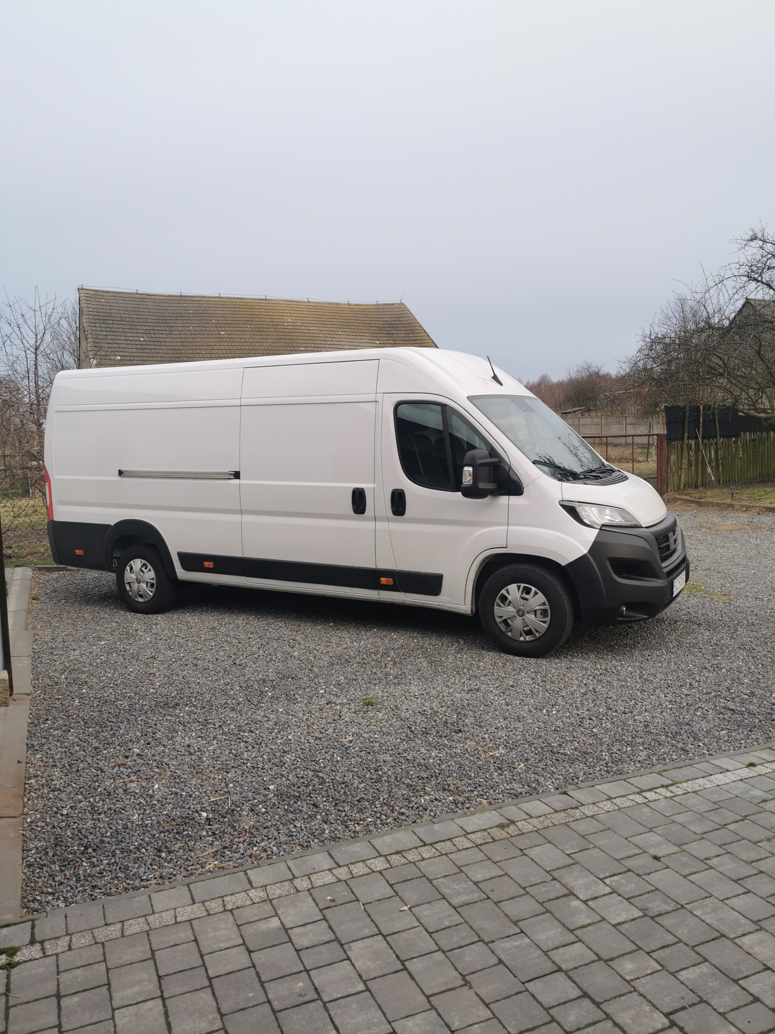 Biały, dostawczy bus zaparkowany na żwirowym podjeździe przed domem z szarym dachem w pochmurny dzień.