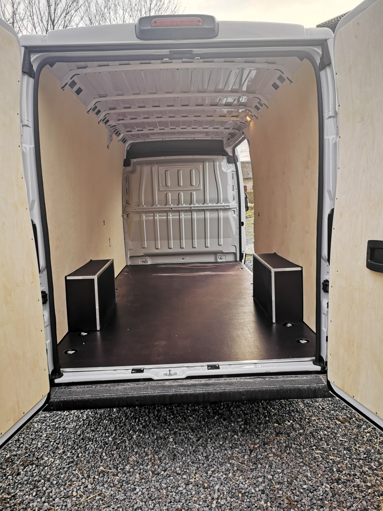 Wnętrze białego busa wyłożone jasną sklejką, z zamontowanymi dwoma czarnymi skrzynkami przy ścianach, widok z zewnątrz przez otwarte tylne drzwi.