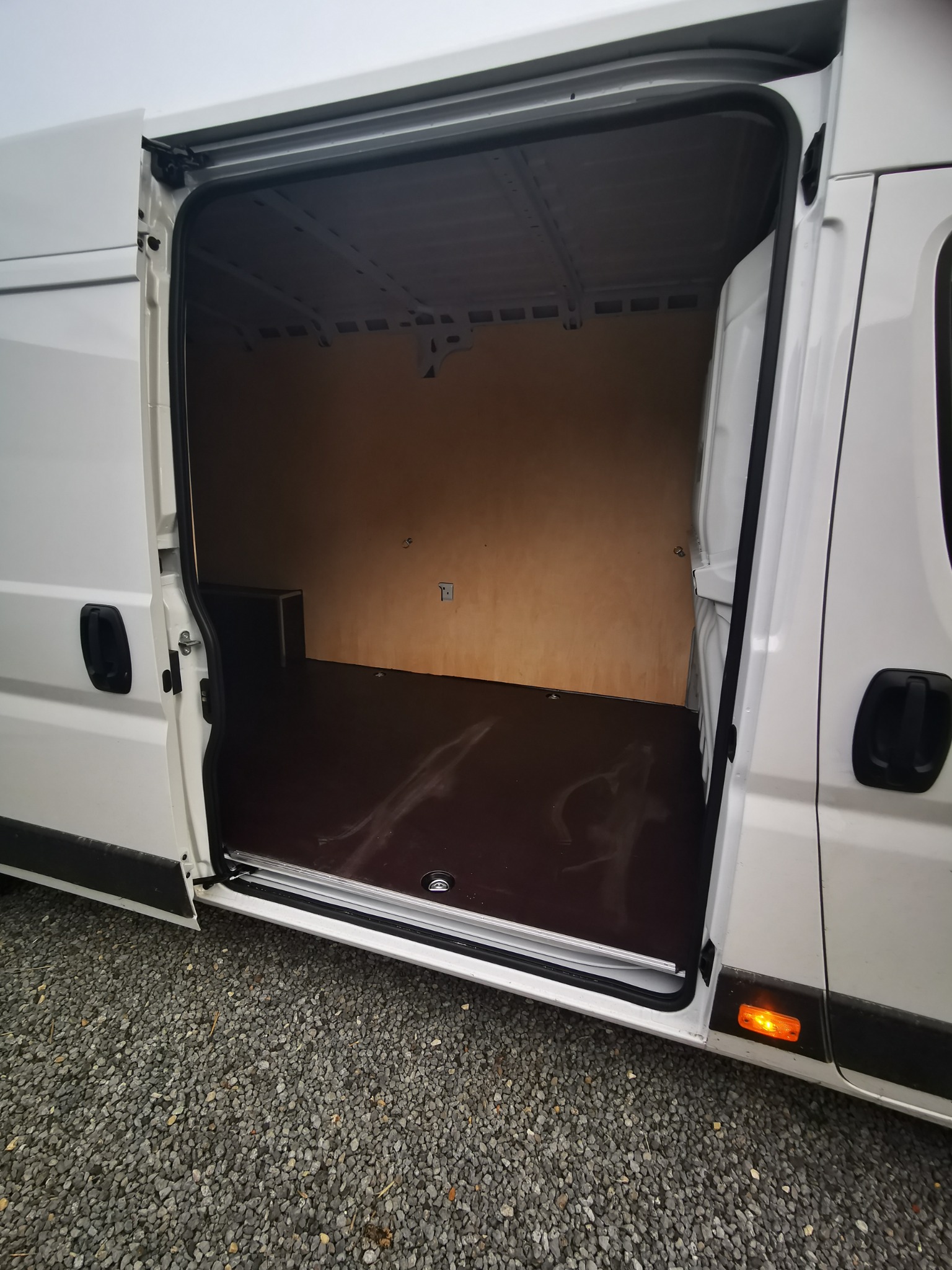 Wnętrze pustego, białego busa z otwartymi drzwiami bocznymi; widoczna drewniana podłoga i ściany boczne przedziału ładunkowego, oraz niewielka skrzynka przy ścianie.