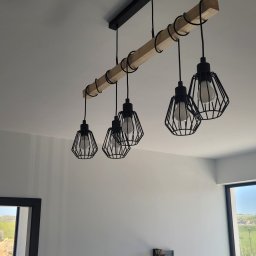 VOLT-RUS Instalacje elektryczne Rafał Rus - Zawieszona pod sufitem lampa z pięcioma żarówkami w czarnych, drucianych kloszach, zamocowana na drewnianej belce z czarnymi pierścieniami, widoczna z dołu, w tle ściana i okno z widokiem...