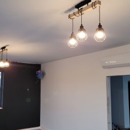 VOLT-RUS Instalacje elektryczne Rafał Rus - Nowoczesna, potrójna lampa wisząca z metalowymi kloszami w kształcie klatek, zamontowana na drewnianej belce w surowym wnętrzu w trakcie remontu, podłoga zabezpieczona folią, widoczna skrzynka...
