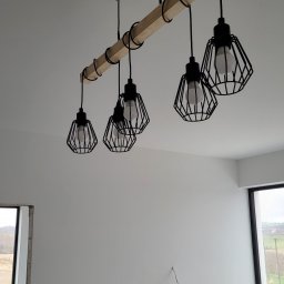 VOLT-RUS Instalacje elektryczne Rafał Rus - Zawieszone, czarne, klatkowe lampy z żarówkami LED zamontowane na drewnianej belce, przymocowanej do sufitu w nowym, jasnym pomieszczeniu z widocznymi przewodami na ścianie.