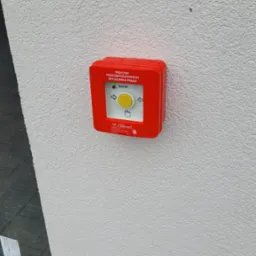 Czerwony, natynkowy przycisk przeciwpożarowy z żółtym guzikiem i napisem 'Dozór', zamontowany na białej, chropowatej ścianie budynku.