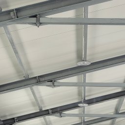 VOLT-RUS Instalacje elektryczne Rafał Rus - Wnętrze hali magazynowej z widoczną konstrukcją stalową dachu i zamontowanymi okrągłymi lampami przemysłowymi LED z metalowymi radiatorami.