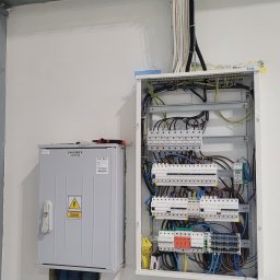 VOLT-RUS Instalacje elektryczne Rafał Rus - Rozdzielnica elektryczna z otwartymi drzwiami, widoczne bezpieczniki, kable, oraz dodatkowa szara skrzynka z symbolem ostrzegawczym napięcia, kable poprowadzone w korytach i peszlach.