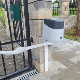 VOLT-RUS Instalacje elektryczne Rafał Rus - Szara automatyka do bramy zamontowana na słupku z szarej kostki, widoczny fragment czarnej bramy, skrzynka na listy i oświetlenie zewnętrzne.