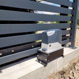 VOLT-RUS Instalacje elektryczne Rafał Rus - Szara, nowoczesna brama przesuwna z automatyką firmy Nice, zamontowana na betonowej podstawie, widoczne wystające kable.