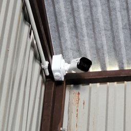 VOLT-RUS Instalacje elektryczne Rafał Rus - Kamera monitoringu zamontowana wewnątrz magazynu w Pogorzanach, biała obudowa kamery przymocowana do metalowej ramy i blachy falistej.