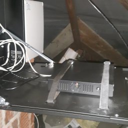 VOLT-RUS Instalacje elektryczne Rafał Rus - Zamontowany w przestrzeni poddasza rejestrator monitoringu wizyjnego w czarnej obudowie, przymocowany pasami do metalowej półki, z widocznymi kablami i fragmentem instalacji elektrycznej.
