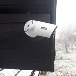 VOLT-RUS Instalacje elektryczne Rafał Rus - Biała kamera BCS zamontowana na ciemnej drewnianej elewacji budynku, widok z góry na ośnieżony trawnik i drzewa we mgle.