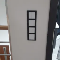 Czarny panel z czterema białymi włącznikami światła zamontowany na białej ścianie, widoczny fragment schod&oacute;w drewnianych i futryna drzwiowa.