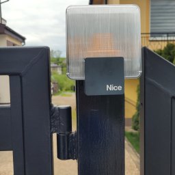 VOLT-RUS Instalacje elektryczne Rafał Rus - Lampa ostrzegawcza marki Nice zamontowana na czarnym, metalowym słupku bramy wjazdowej, z widocznym tłem zabudowy jednorodzinnej.