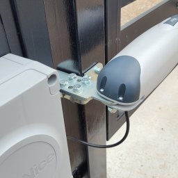 VOLT-RUS Instalacje elektryczne Rafał Rus - Szczegółowe ujęcie zamontowanego napędu elektrycznego bramy NICE, z widocznymi śrubami mocującymi i kablem zasilającym, na tle czarnej metalowej konstrukcji bramy.
