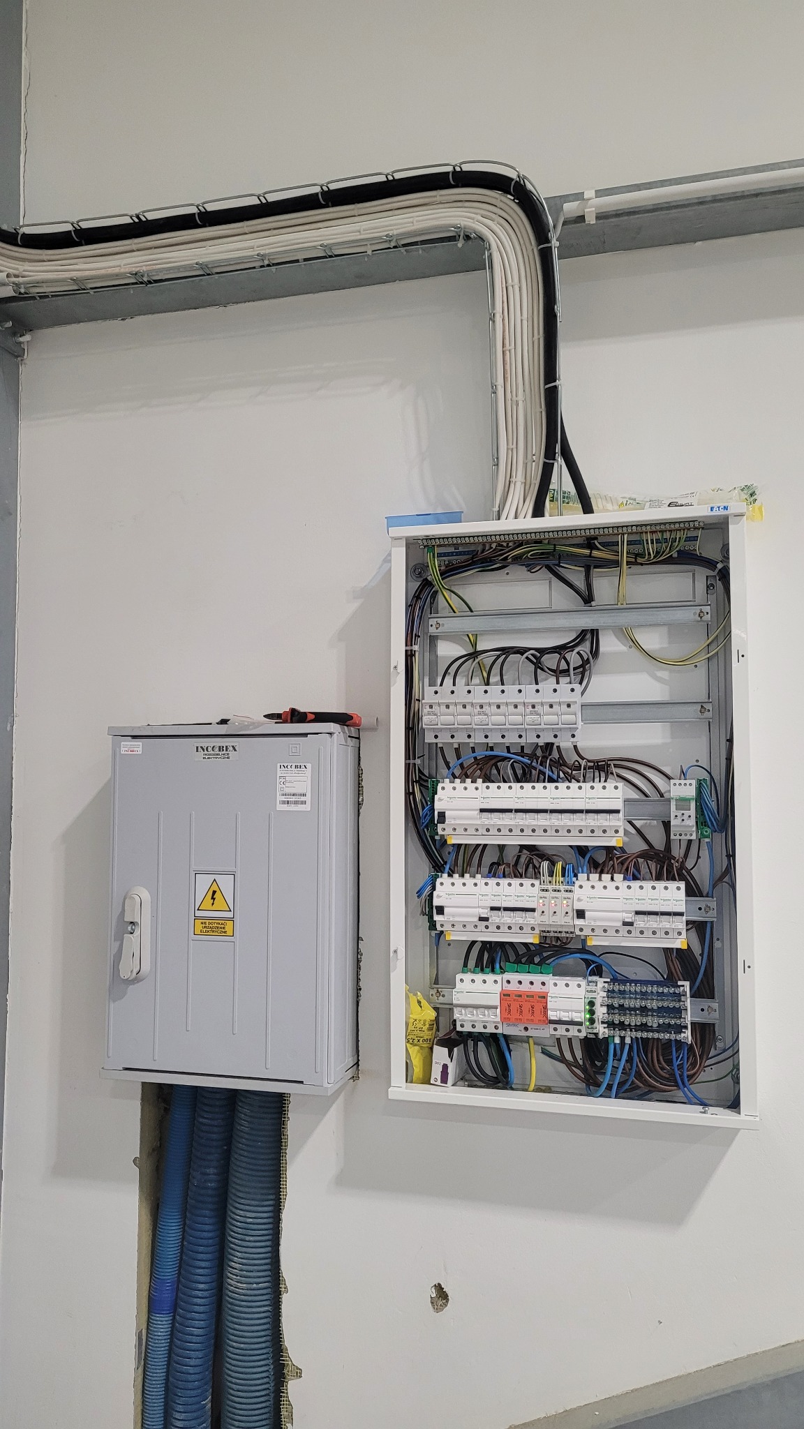 Rozdzielnica elektryczna z otwartymi drzwiami, widoczne bezpieczniki, kable, oraz dodatkowa szara skrzynka z symbolem ostrzegawczym napięcia, kable poprowadzone w korytach i peszlach.