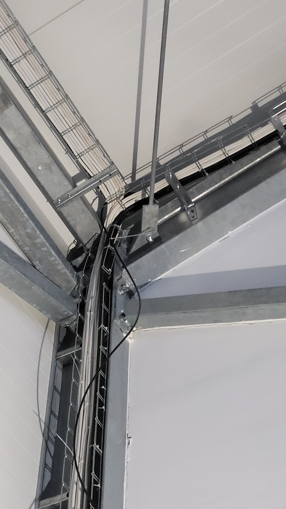 Metalowe koryta kablowe z przewodami ułożone pod sufitem, widoczne mocowania do konstrukcji stalowej, zbliżenie na zakręt kabli w narożniku.