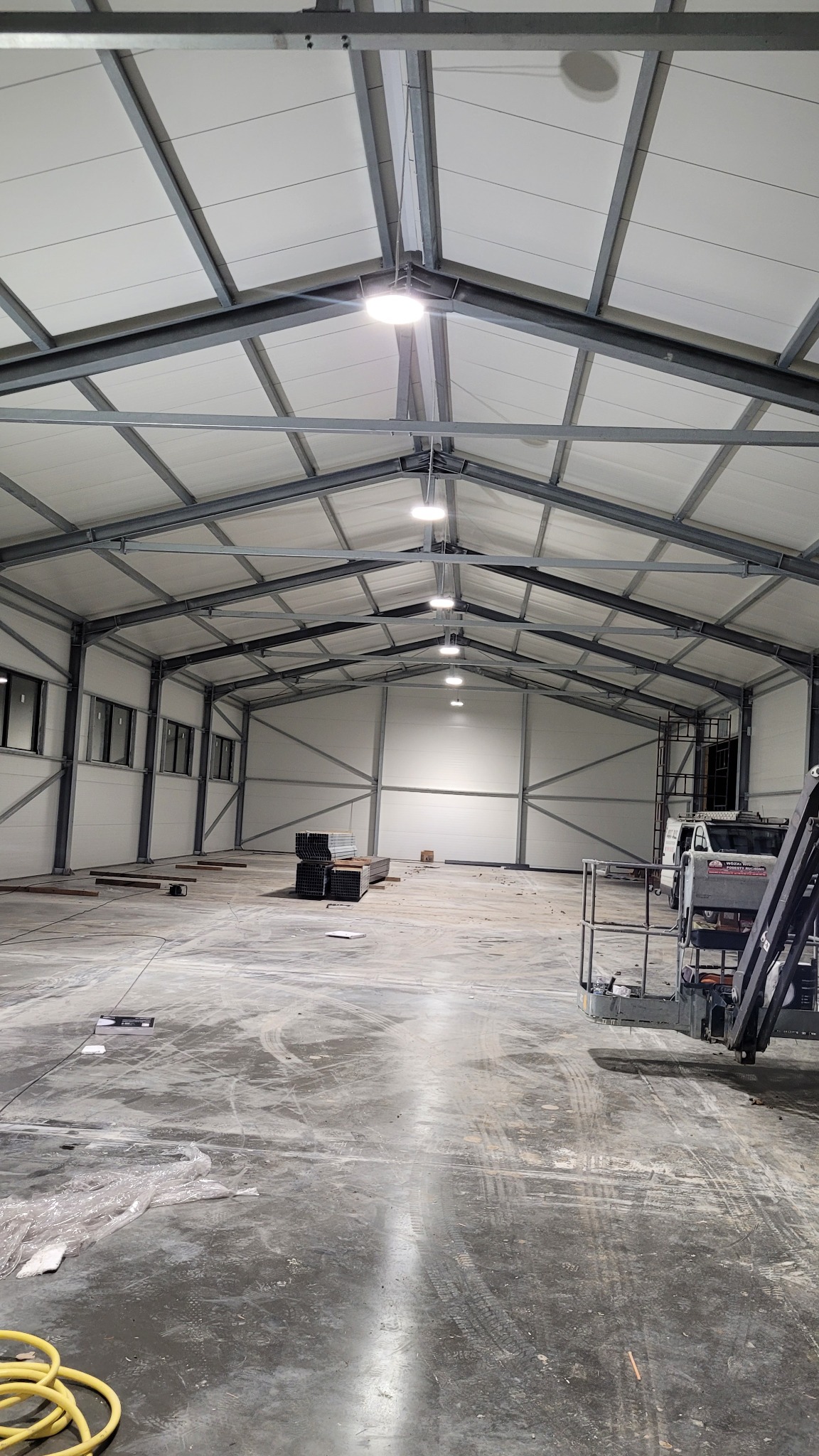 Wnętrze hali magazynowej z widoczną konstrukcją stalową dachu, oświetleniem LED, fragmentami materiałów budowlanych i sprzętu, ukazujące etap prac wykończeniowych.