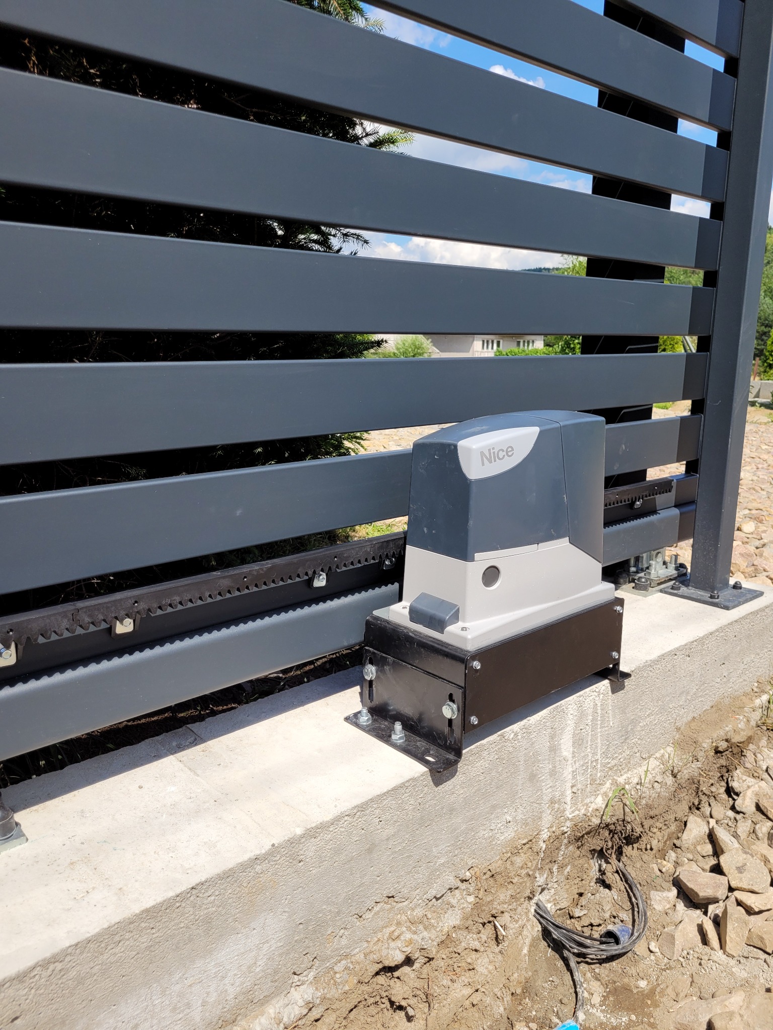 Szara, nowoczesna brama przesuwna z automatyką firmy Nice, zamontowana na betonowej podstawie, widoczne wystające kable.