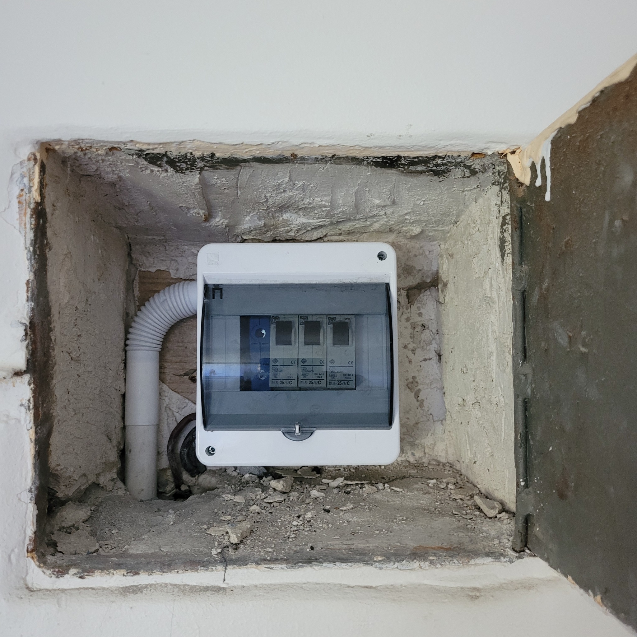 Zamknięta skrzynka bezpiecznikowa z widocznymi bezpiecznikami, umieszczona we wnęce w ścianie, z widoczną rurą osłonową przewodów.