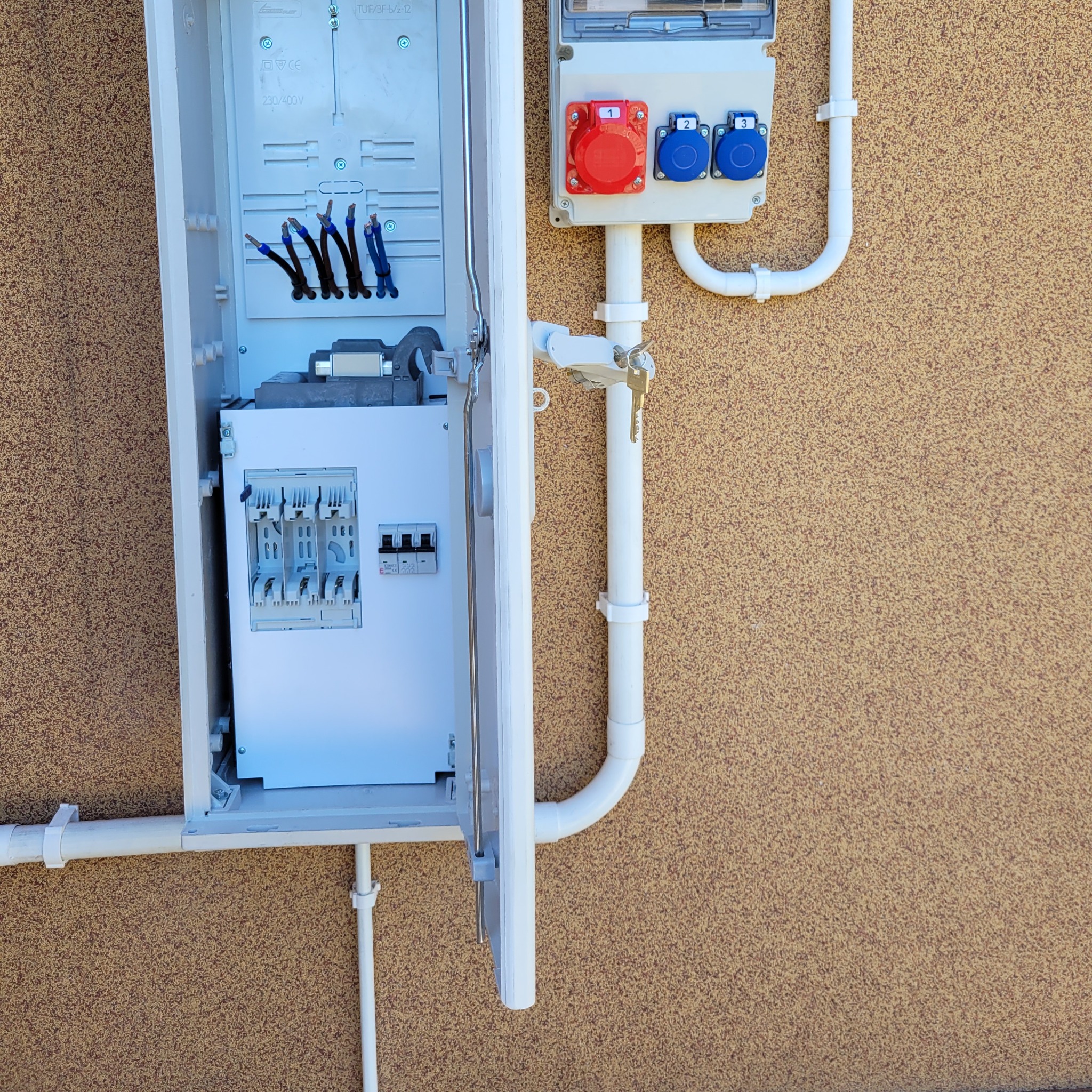 Otwarta skrzynka elektryczna z widocznymi przewodami, bezpiecznikami i gniazdami siłowymi na zewnątrz budynku o beżowej elewacji.