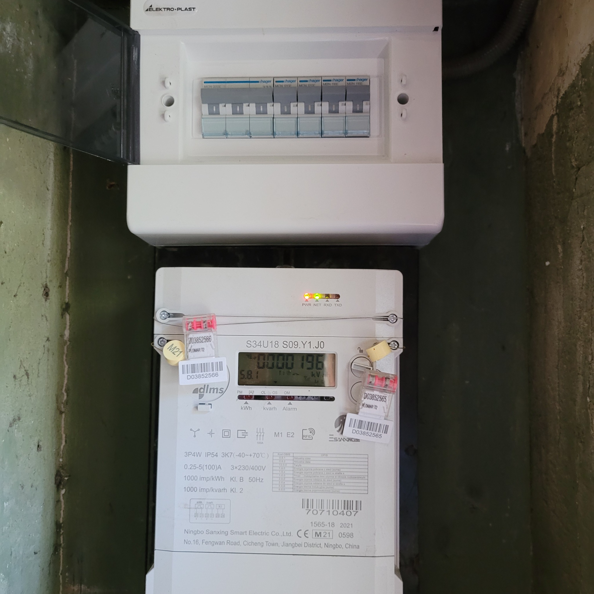 Skrzynka rozdzielcza Elektro-Plast z widocznymi bezpiecznikami Hager oraz licznik energii elektrycznej Ningbo Sanxing z plombami, zamontowane na starej, pomalowanej ścianie.