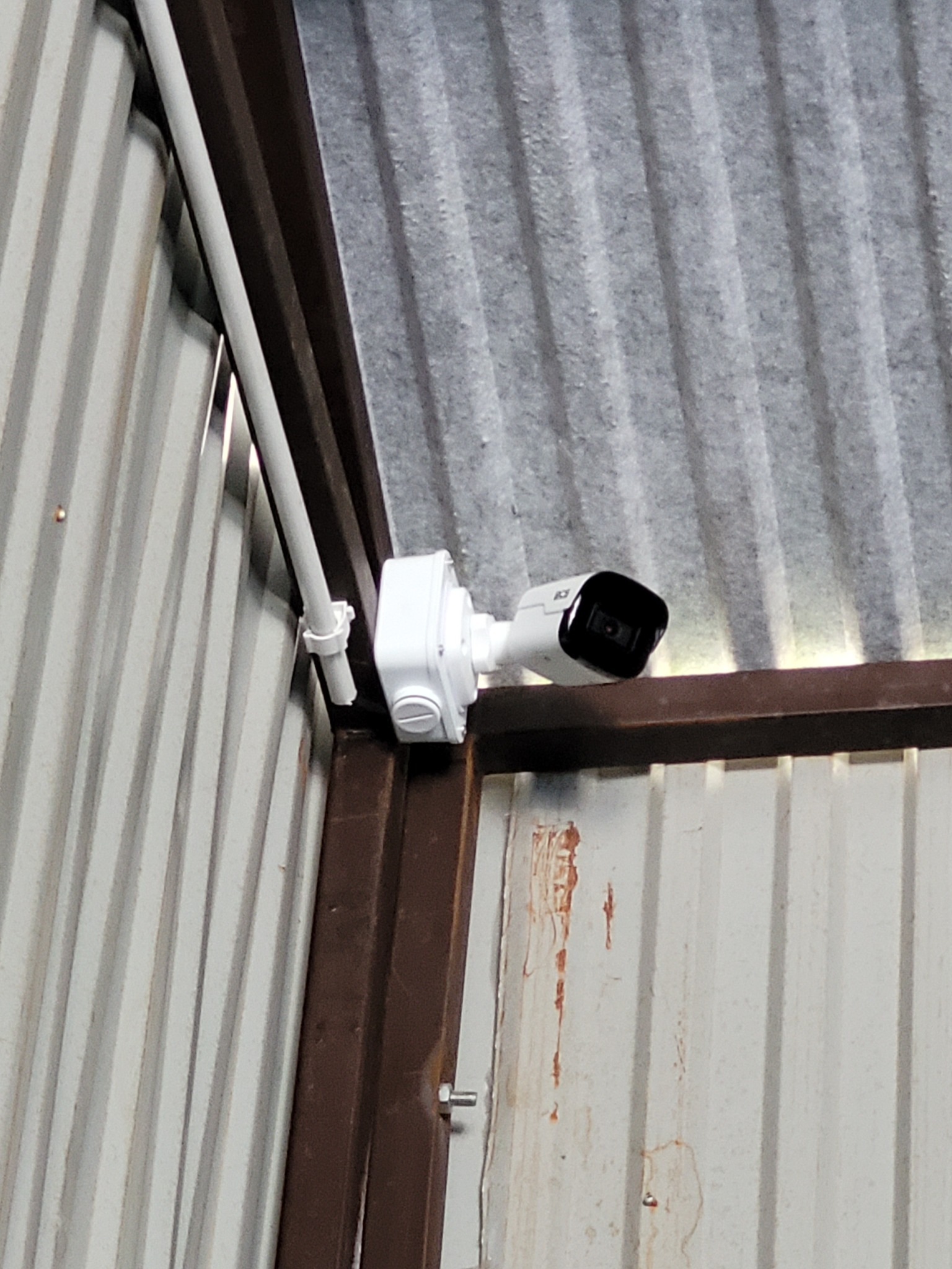 Kamera monitoringu zamontowana wewnątrz magazynu w Pogorzanach, biała obudowa kamery przymocowana do metalowej ramy i blachy falistej.