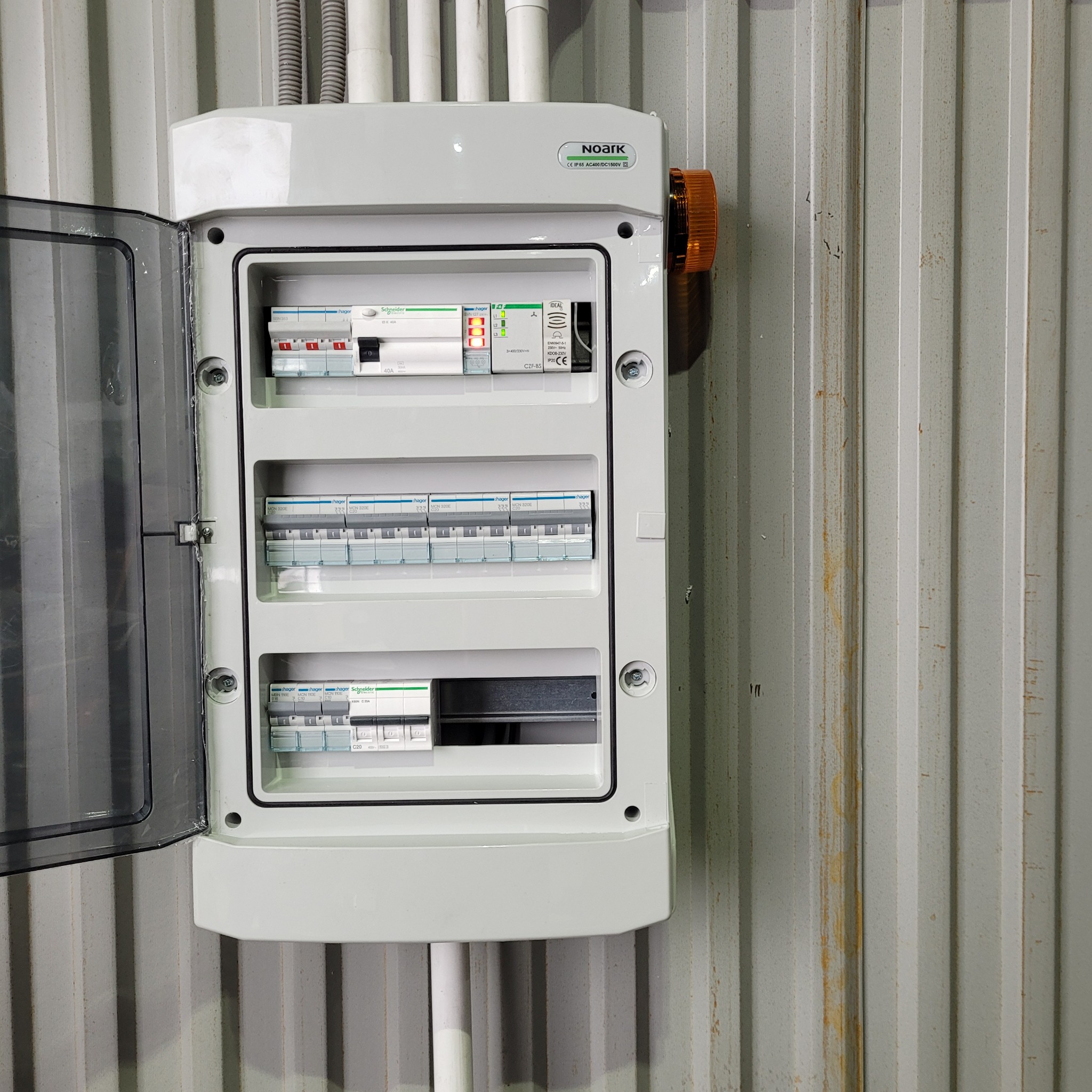 Otwarta, biała skrzynka rozdzielcza Noark z widocznymi bezpiecznikami Hager i Schneider, zamontowana na szarej, blaszanej ścianie z widoczną lampką sygnalizacyjną po prawej stronie.