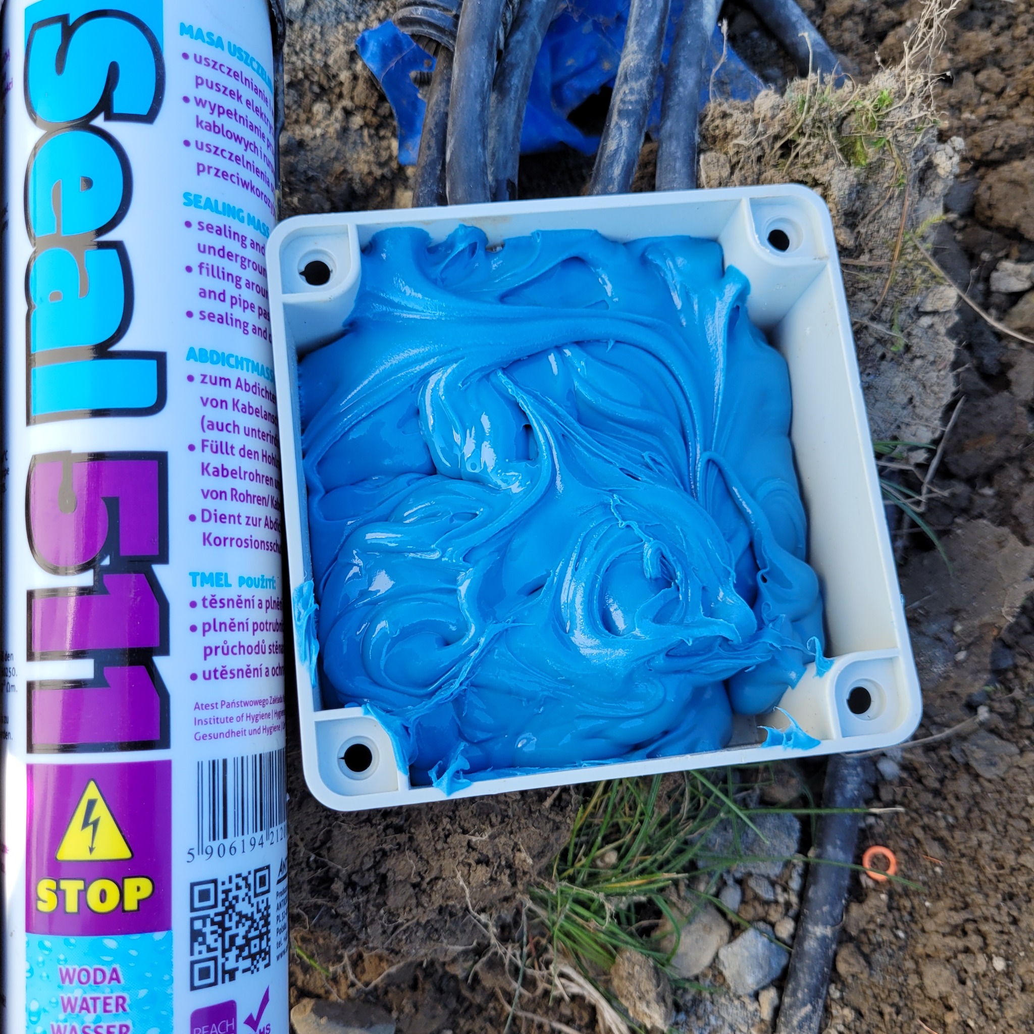 Niebieska masa uszczelniająca w białym pudełku, obok puszka z opisem w kilku językach, widoczne kable i ziemia.