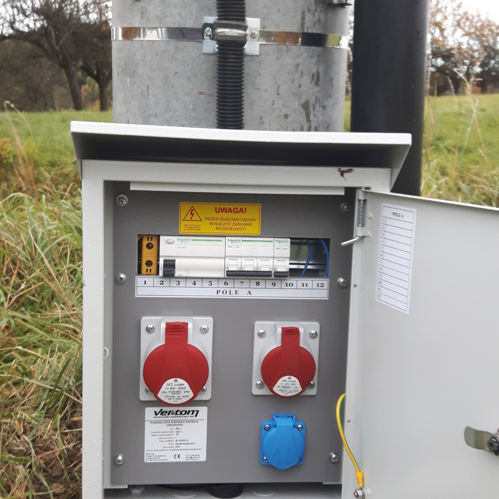 Otwarta skrzynka elektryczna zamontowana na słupie, z widocznymi bezpiecznikami, gniazdami siłowymi i ostrzeżeniem o wyłączeniu zasilania przed zdjęciem osłony, umieszczona na zewnątrz w otoczeniu...