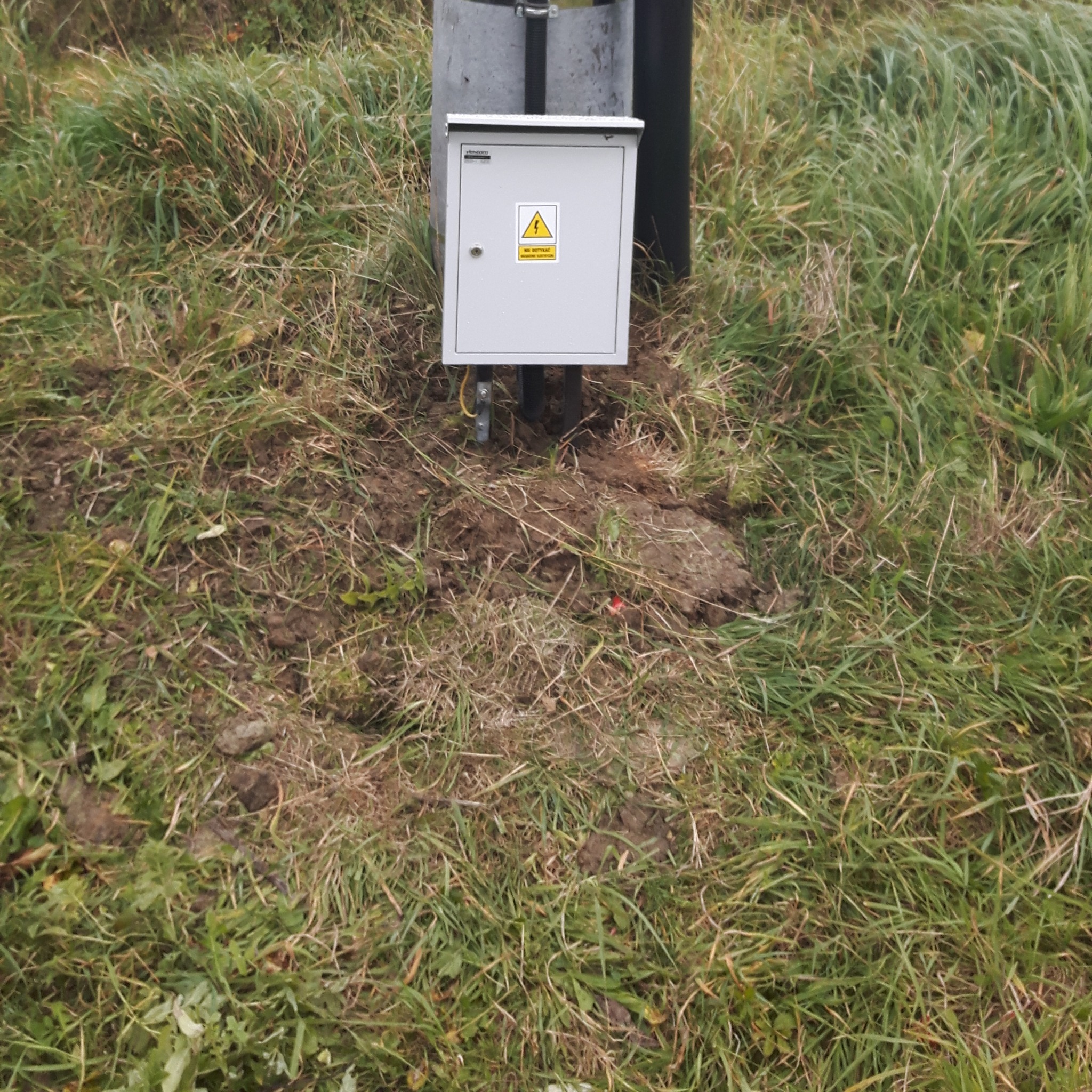 Szara skrzynka elektryczna z żółtym znakiem ostrzegawczym, zamontowana na słupku w trawie.
