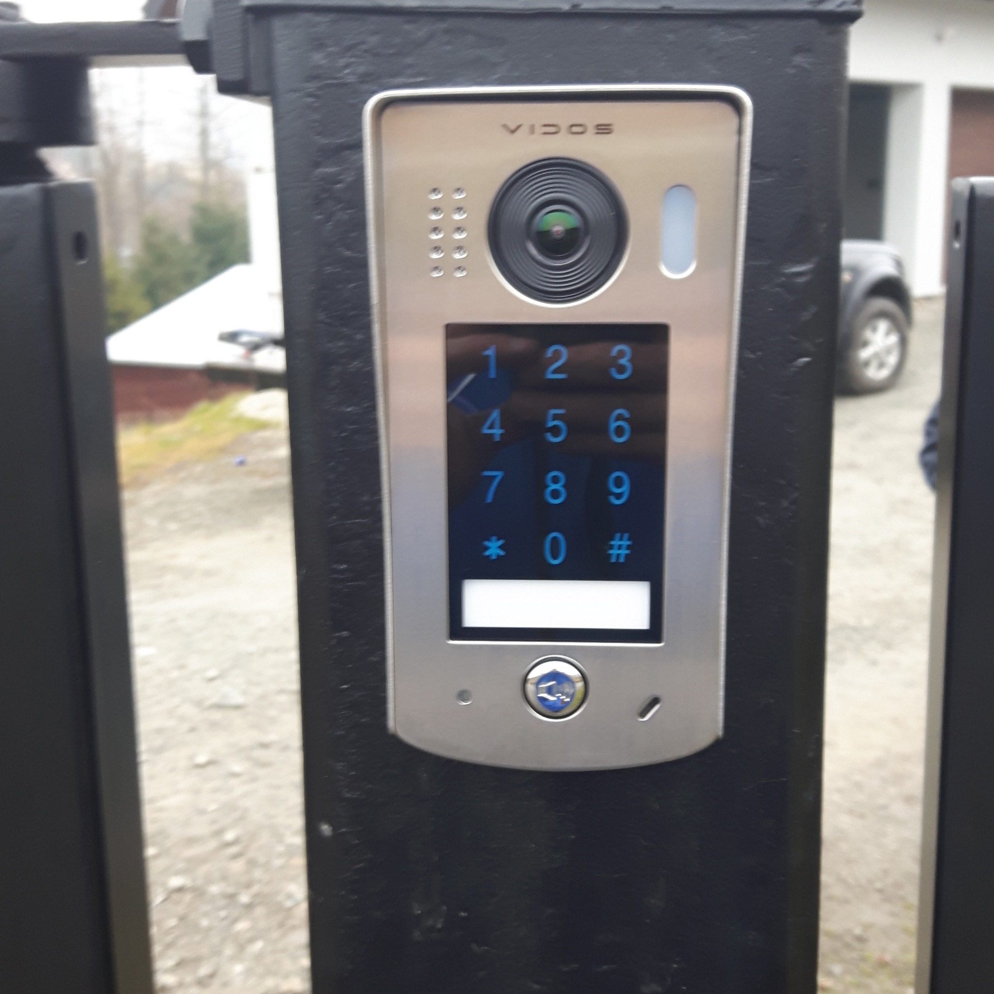 Panel domofonu Vidos z klawiaturą numeryczną i kamerą zamontowany na czarnym słupku bramy wjazdowej, widoczny fragment samochodu i otoczenie posesji.