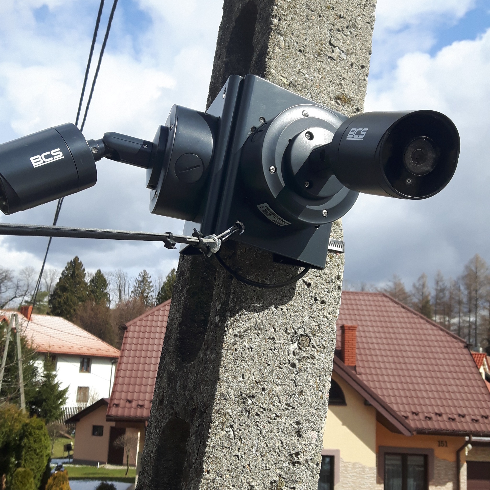 Dwie czarne kamery monitoringu z logo BCS zamontowane na betonowym słupie energetycznym, w tle domy jednorodzinne z brązowymi dachami i pochmurne niebo.