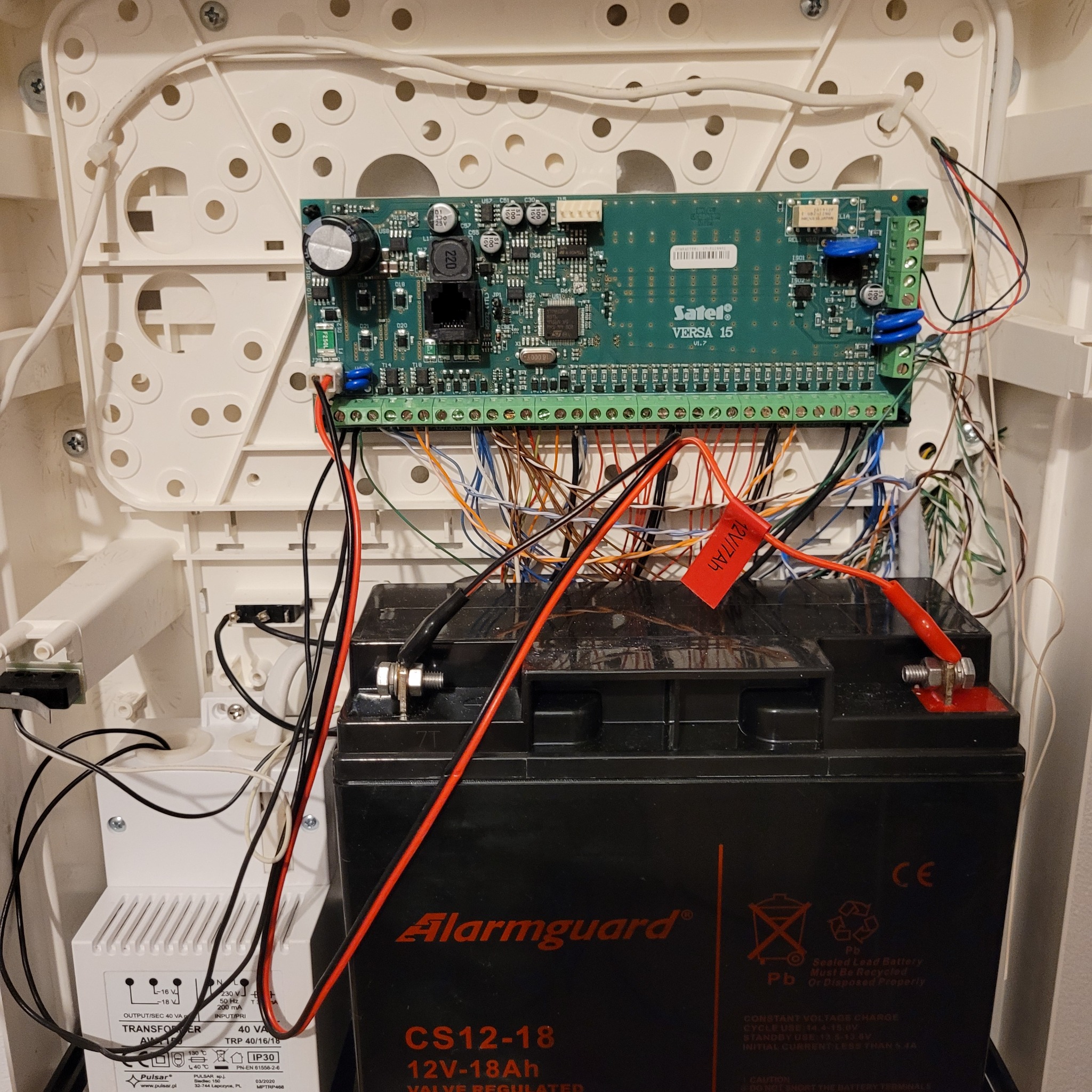Centrala alarmowa Satel Versa 15 w białej obudowie z podłączonym akumulatorem Alarmguard i transformatorem Pulsar, widoczne liczne przewody połączeniowe.