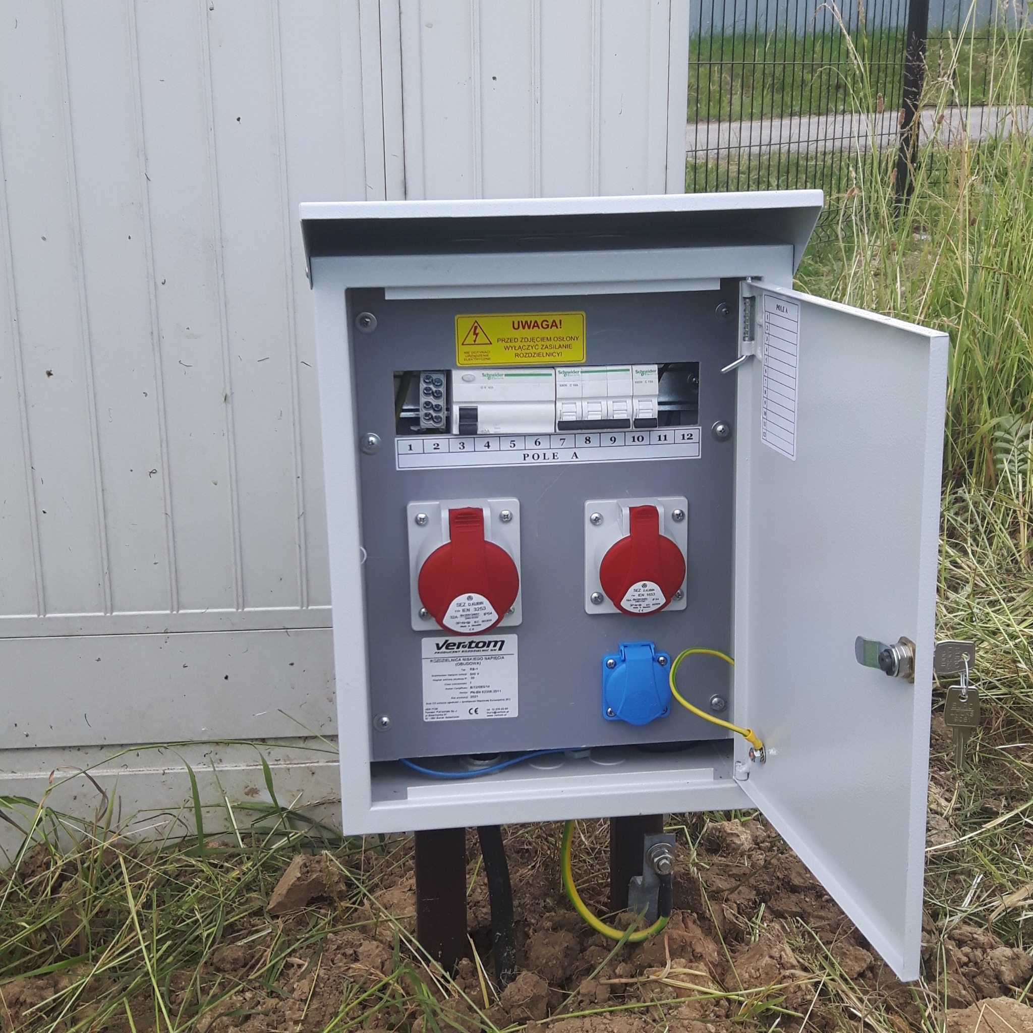 Otwarta skrzynka elektryczna z gniazdami przemysłowymi, bezpiecznikami i naklejką ostrzegawczą, zamontowana na zewnątrz, widoczne przewody i uziemienie.