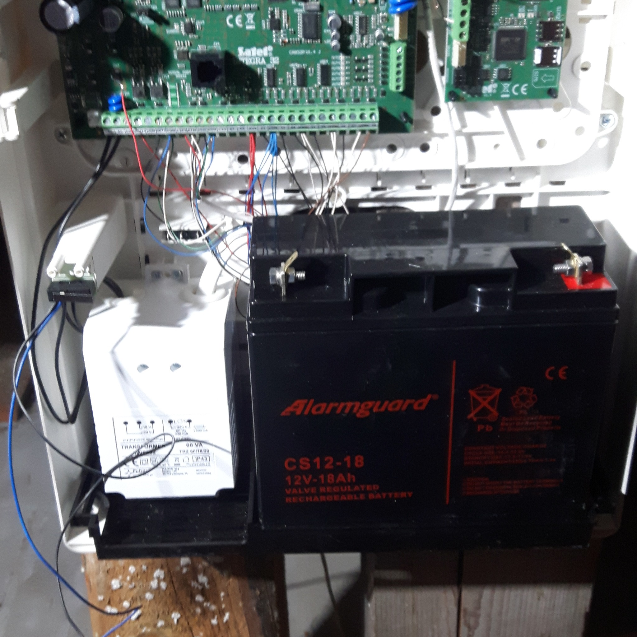 Panel sterowania alarmu z podłączonymi przewodami i akumulatorem Alarmguard CS12-18, widoczny transformator Pulsar TRZ 60/18/20.