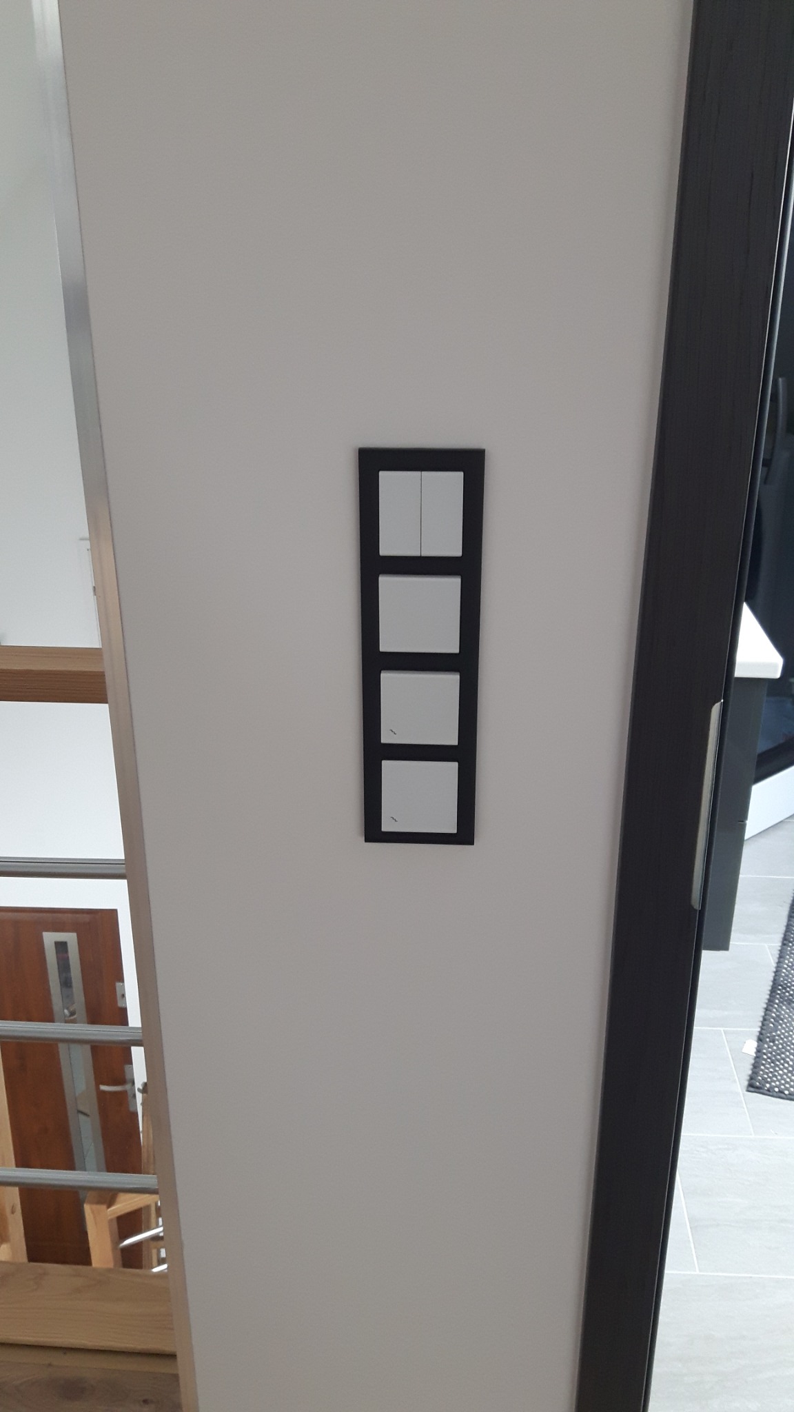 Czarny panel z czterema białymi włącznikami światła zamontowany na białej ścianie, widoczny fragment schodów drewnianych i futryna drzwiowa.