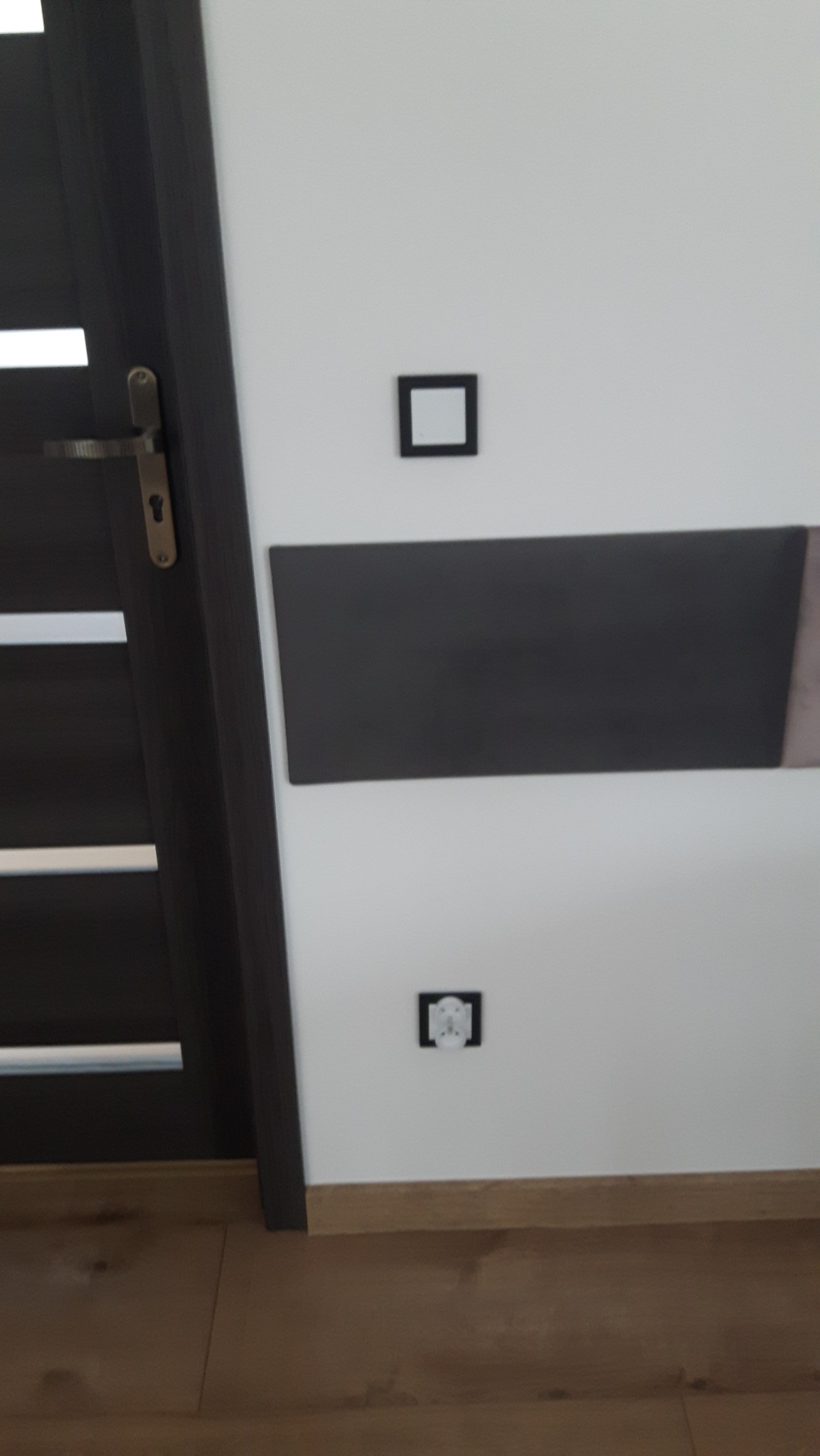 Biała ściana z czarnymi elementami dekoracyjnymi: kwadratowa ramka, prostokątny panel i gniazdko elektryczne z czarną obwódką, widoczny fragment ciemnych drzwi z klamką.