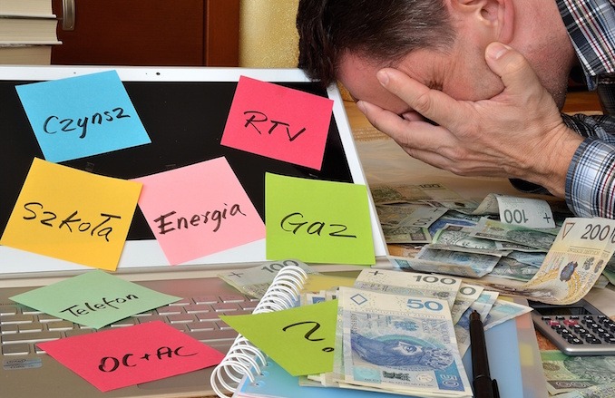 Zrozpaczony mężczyzna zakrywa twarz dłońmi, otoczony stosem banknotów w polskich złotych, laptopem z naklejonymi kolorowymi karteczkami z napisanymi opłatami: Czynsz, RTV, Szkoła, Energia, Gaz...