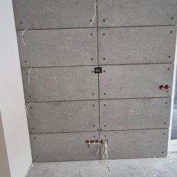 Damian Turcza Dampro - Ściana wykończona panelami imitującymi beton, z widocznymi otworami na gniazdka elektryczne i wystającymi kablami, surowy betonowy wygląd w trakcie prac wykończeniowych.