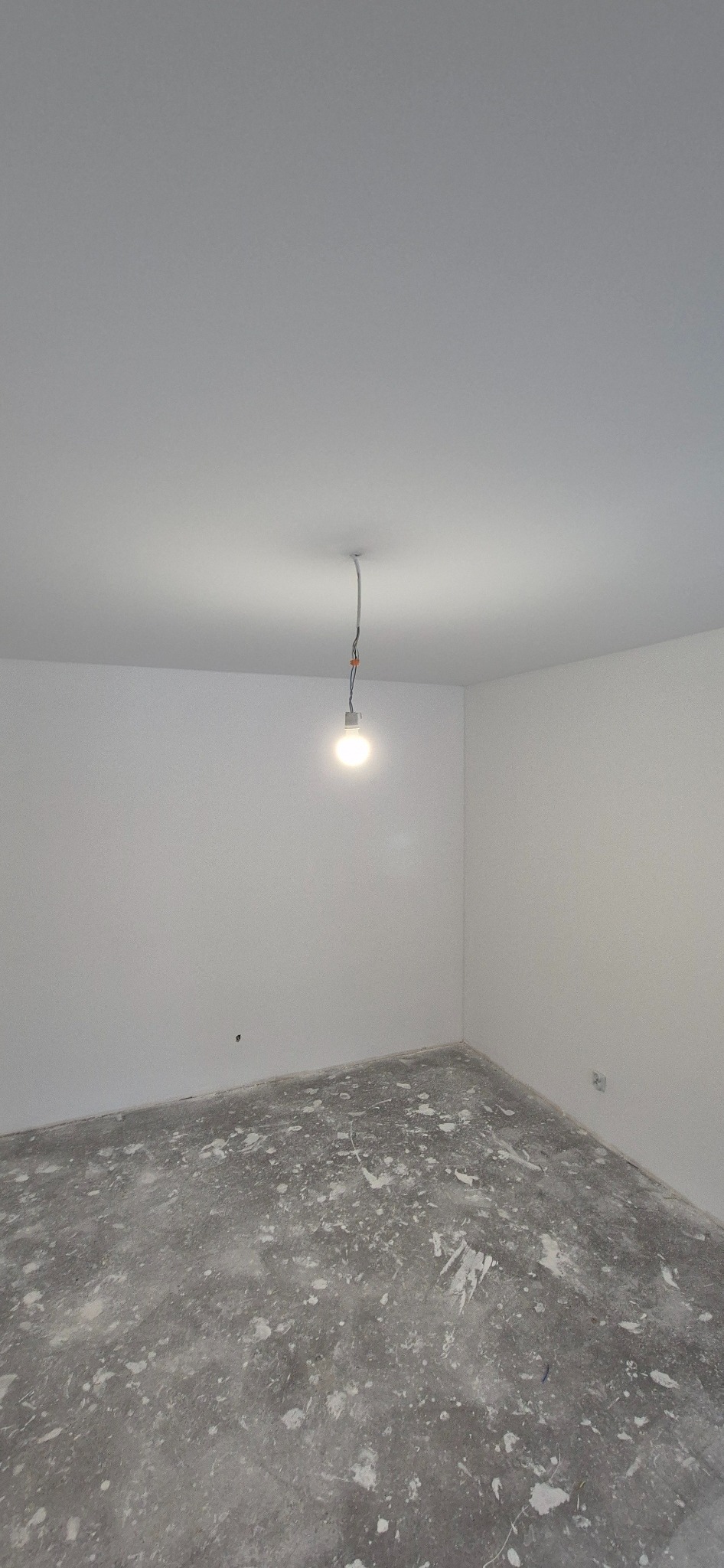 Pokój w trakcie remontu z surowymi, białymi ścianami i podłogą pokrytą resztkami farby lub gipsu, oświetlony wiszącą żarówką.