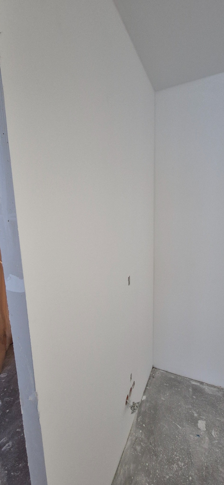 Wnętrze pokoju w trakcie remontu, ściany pokryte świeżą, jasną warstwą wykończeniową, widoczne niedoskonałości i pozostałości po instalacji elektrycznej przy podłodze, surowy beton na podłodze.