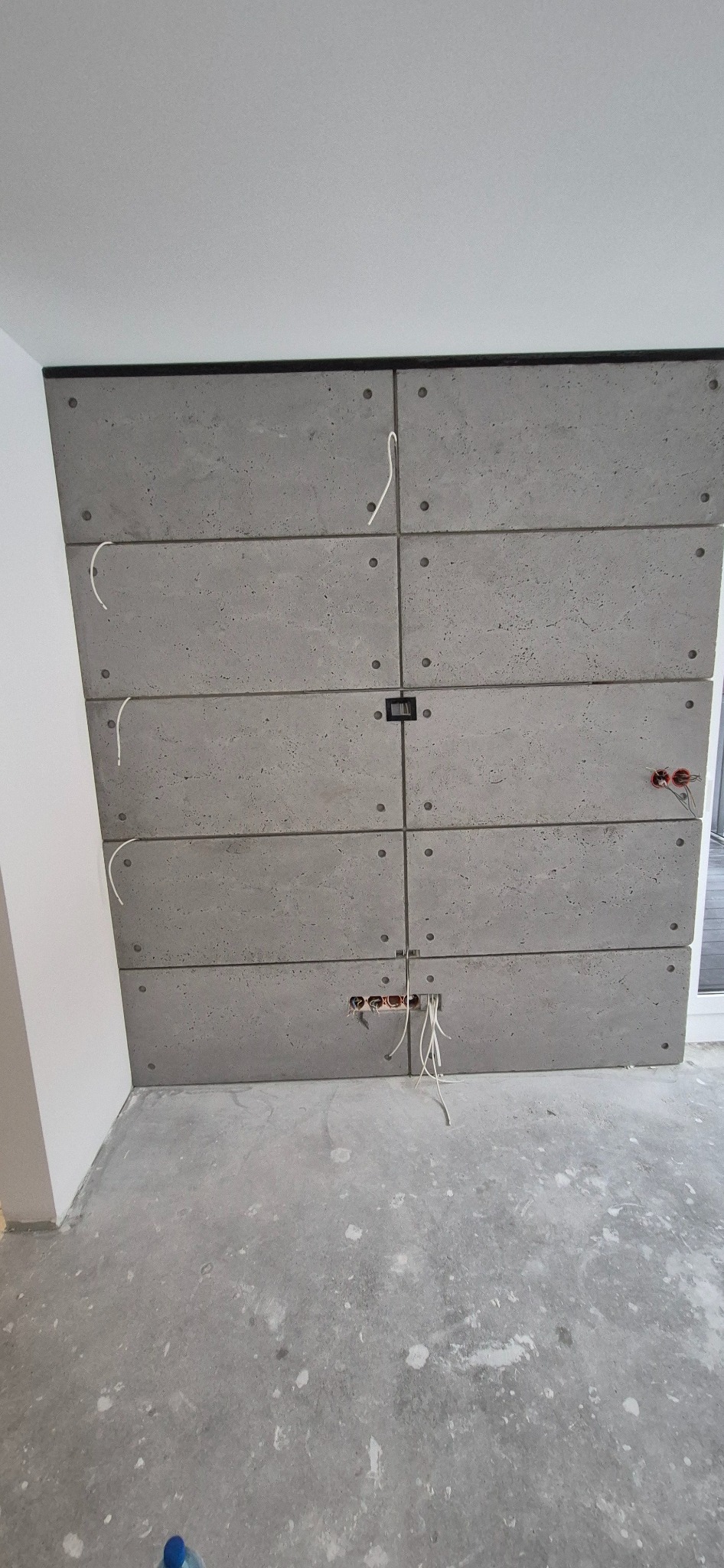 Ściana wykończona panelami imitującymi beton, z widocznymi otworami na gniazdka elektryczne i wystającymi kablami, surowy betonowy wygląd w trakcie prac wykończeniowych.